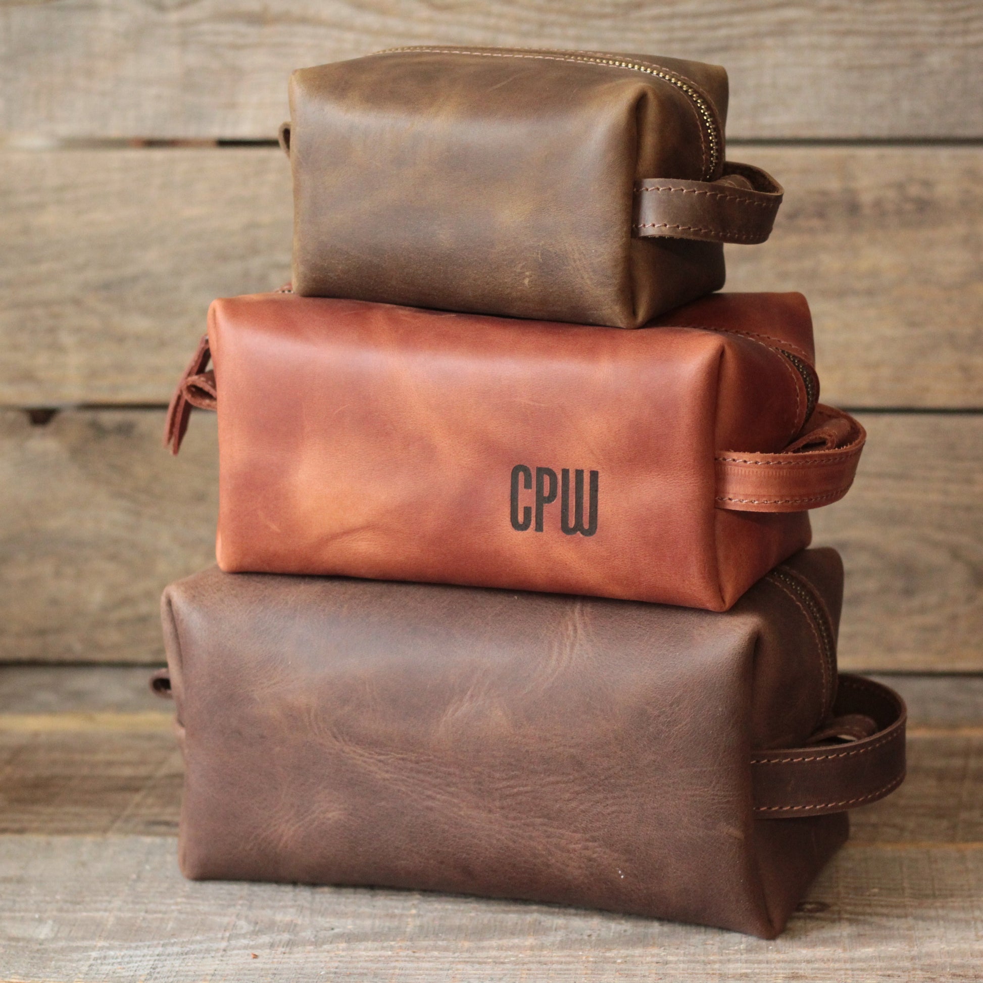  Trzy skórzane kosmetyczki podróżne w rozmiarach Mini, Duży i Bardzo duży ustawione obok siebie na drewnianym tle.