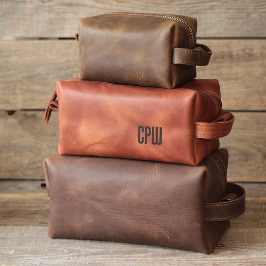  Trzy skórzane kosmetyczki podróżne w rozmiarach Mini, Duży i Bardzo duży ustawione obok siebie na drewnianym tle.